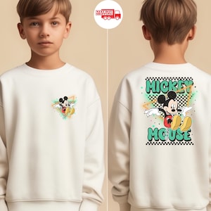May include: Cream-colored sweatshirt featuring Mickey Mouse. The front has a small graphic of Mickey in a colorful triangle. The back displays a larger design with Mickey, the words "Mickey Mouse," and a checkered pattern.