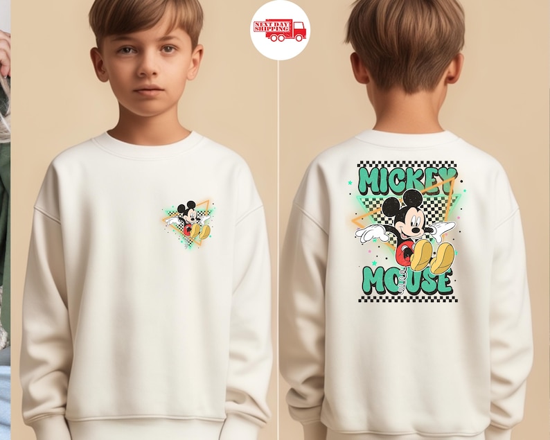 May include: Cream-colored sweatshirt featuring Mickey Mouse. The front has a small graphic of Mickey in a colorful triangle. The back displays a larger design with Mickey, the words "Mickey Mouse," and a checkered pattern.