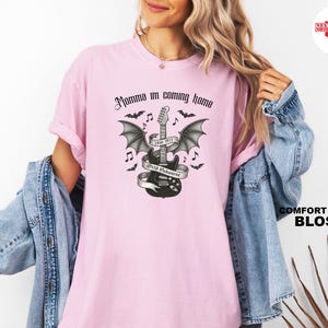 Puede incluir: Una camiseta rosa claro con la frase "Momma I'm coming home" en fuente g&oacute;tica. Un gr&aacute;fico en blanco y negro de una guitarra con alas de murci&eacute;lago, notas musicales y murci&eacute;lagos est&aacute; centrado en la camiseta. La camiseta es un Comfort Colors Blossom.