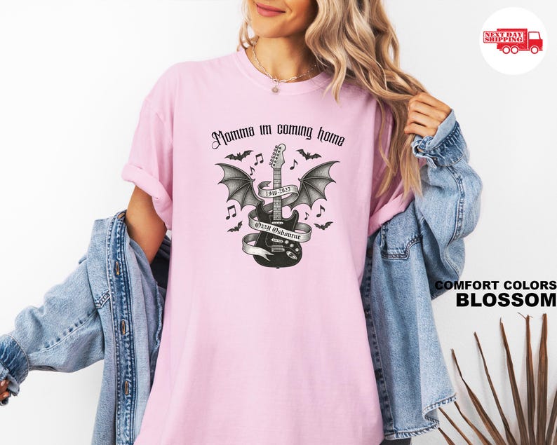 Puede incluir: Una camiseta rosa claro con la frase "Momma I'm coming home" en fuente g&oacute;tica. Un gr&aacute;fico en blanco y negro de una guitarra con alas de murci&eacute;lago, notas musicales y murci&eacute;lagos est&aacute; centrado en la camiseta. La camiseta es un Comfort Colors Blossom.