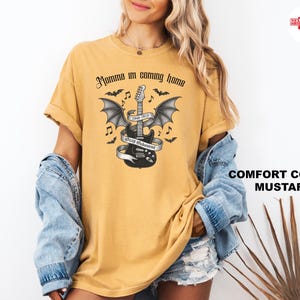 Puede incluir: Camiseta de color mostaza con una guitarra negra con alas de murci&eacute;lago, una pancarta y el texto "Momma I'm coming home". El dise&ntilde;o incluye murci&eacute;lagos y notas musicales. La camiseta es de color c&oacute;modo.