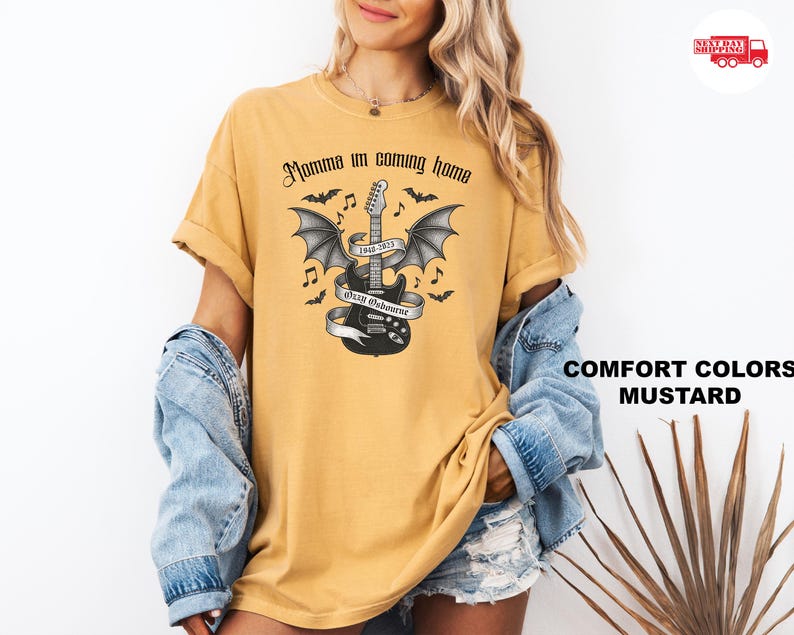 Puede incluir: Camiseta de color mostaza con una guitarra negra con alas de murci&eacute;lago, una pancarta y el texto "Momma I'm coming home". El dise&ntilde;o incluye murci&eacute;lagos y notas musicales. La camiseta es de color c&oacute;modo.