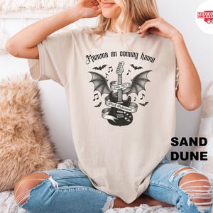 Puede incluir: Camiseta color arena con un dise&ntilde;o gr&aacute;fico en blanco y negro de una guitarra con alas de murci&eacute;lago, notas musicales y el texto "Momma I'm coming home" y "Ozzy Osbourne 1948-2023".