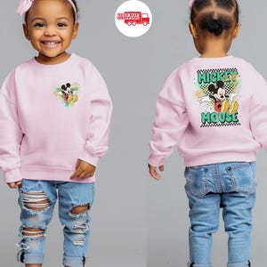 May include: A light pink sweatshirt featuring a Mickey Mouse graphic on the front and back. The front graphic is a small Mickey Mouse design with colorful accents. The back of the sweatshirt has a larger Mickey Mouse graphic with the words "Mickey Mouse".