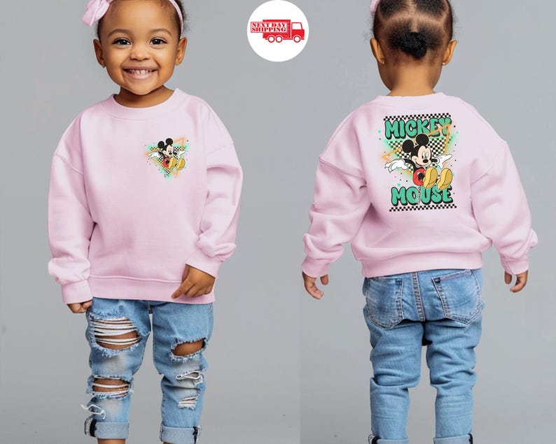 May include: A light pink sweatshirt featuring a Mickey Mouse graphic on the front and back. The front graphic is a small Mickey Mouse design with colorful accents. The back of the sweatshirt has a larger Mickey Mouse graphic with the words "Mickey Mouse".