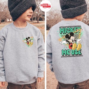 May include: A gray sweatshirt featuring Mickey Mouse. The front has a small graphic of Mickey, while the back displays a larger design with "Mickey Mouse" in green lettering. The design includes a checkered pattern and colorful accents.