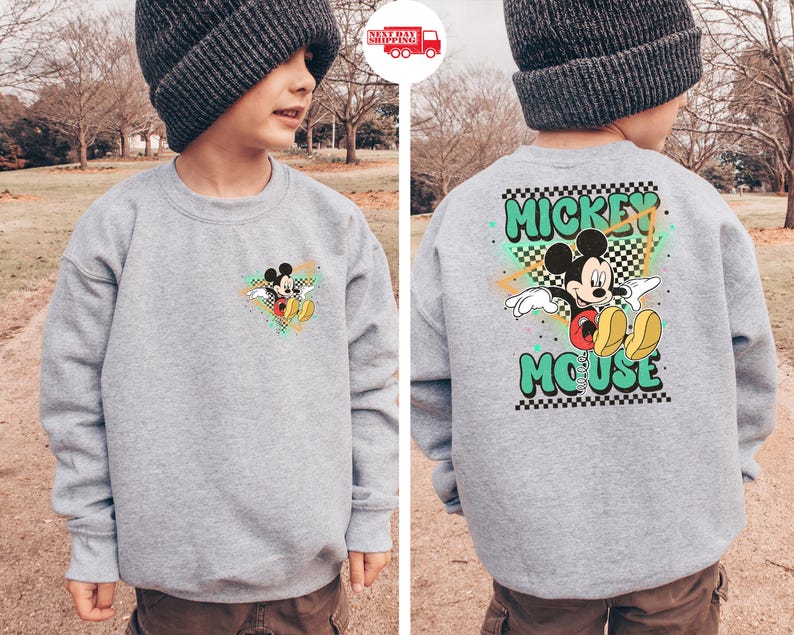 May include: A gray sweatshirt featuring Mickey Mouse. The front has a small graphic of Mickey, while the back displays a larger design with "Mickey Mouse" in green lettering. The design includes a checkered pattern and colorful accents.