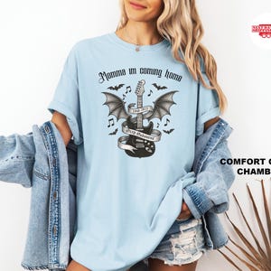 Puede incluir: Camiseta azul claro con un gr&aacute;fico en blanco y negro de una guitarra con alas de murci&eacute;lago, notas musicales y el texto "Momma I'm coming home" y "Ozzy Osbourne". La camiseta est&aacute; hecha de un material suave y c&oacute;modo.