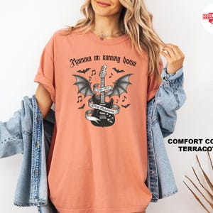 Puede incluir: Camiseta color terracota con un dise&ntilde;o en blanco y negro de una guitarra con alas de murci&eacute;lago, notas musicales y el texto "Momma I'm coming home". La camiseta tambi&eacute;n tiene el texto "Ozzy Osbourne 1948-2023".
