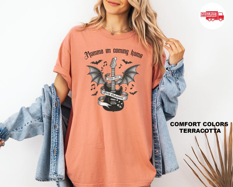 Puede incluir: Camiseta color terracota con un dise&ntilde;o en blanco y negro de una guitarra con alas de murci&eacute;lago, notas musicales y el texto "Momma I'm coming home". La camiseta tambi&eacute;n tiene el texto "Ozzy Osbourne 1948-2023".