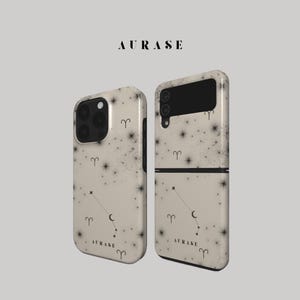 May include: Two phone cases with a cream-colored background and a starry night design. The cases feature black constellations, crescent moons, and the word "AURASE" in black. One case is for a standard phone, and the other is a foldable phone.