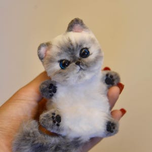 Puede incluir: Una figura de gato persa de peluche gris y blanco, con ojos azules y almohadillas negras. El gato tiene una cola esponjosa y se sostiene en una mano. Un artículo artesanal suave y detallado.