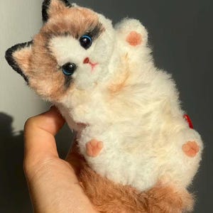 Puede incluir: Un peluche de gato con un cuerpo crema, marcas marrones y una larga cola marrón. El gato tiene ojos azules y almohadillas rosadas. El juguete está diseñado para parecerse a un gato Ragdoll.