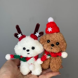 Może przedstawiać: Dwa pluszowe pieski w świątecznych strojach. Jeden biały z porożem renifera i czerwono-zielonym szalikiem. Drugi brązowy, w czerwonej czapce ze śnieżynką i pasującym szaliku. Obydwa są małe i puszyste.