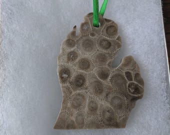 以下が含まれることがあります： ミシガン州の形をした、ベージュと茶色のペトスキーストーンのオーナメント。表面には、化石化したサンゴによる独特の自然な放射状の模様が見られます。上部の穴には、吊り下げるための鮮やかな緑色のリボンが通されています。