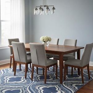 Puede incluir: Un juego de comedor con una mesa rectangular de madera y seis sillas grises tapizadas. La mesa está sobre una alfombra estampada azul y blanca. Una lámpara de araña cuelga sobre la mesa y un jarrón con flores blancas está sobre la mesa.