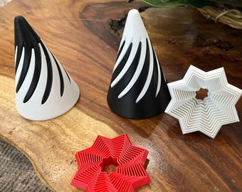 Puede incluir: Una colección de formas geométricas impresas en 3D. Dos objetos en forma de cono, uno negro y otro blanco, presentan rayas blancas y negras curvas. También son visibles dos objetos en forma de estrella, uno rojo y otro blanco. Los objetos se muestran sobre una superficie de madera.