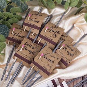 May include: Brown paper wrapped sparklers with personalised labels for wedding favours. The labels feature a heart, the couple's names, and the wedding date. Some labels say "Let Sparks Fly" and "Light Up The Night".