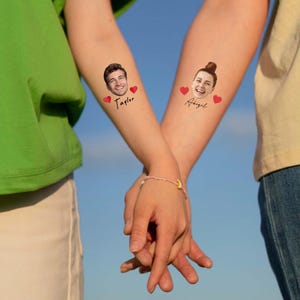May include: Two temporary tattoos applied to forearms. One tattoo features a portrait of a man with the name Taylor and two red hearts. The other tattoo features a portrait of a woman with the name Abigail and two red hearts. The tattoos are black ink.