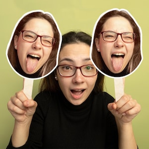 Peut inclure: Une personne tient deux masques avec son propre visage, chacun affichant une expression ludique. La personne porte des lunettes et un col roulé noir, sur un fond vert clair.