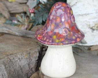May include: A ceramic mushroom figurine with a cream-colored stem and a speckled cap in shades of purple, orange, and yellow. The mushroom is displayed on a rustic wooden surface, evoking a whimsical, natural theme.