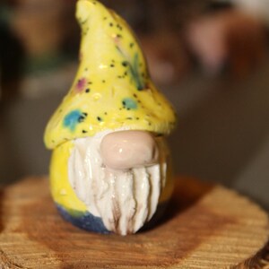 May include: A small ceramic gnome figurine with a yellow hat with black and white polka dots, a white beard, and a pink nose. The gnome is sitting on a wooden log.