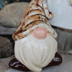 May include: A ceramic garden gnome figurine. The gnome has a cream-colored body and beard, a pink nose, and a brown and cream-colored hat. The gnome is sitting on a wooden surface.