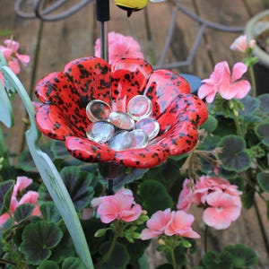 May include: A red and black flower-shaped bird bath filled with clear glass stones, surrounded by pink flowers and green foliage. A decorative garden accent.