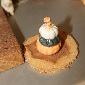 May include: A stack of three miniature ceramic pumpkins, one white, one green, and one orange, on a wooden slice.