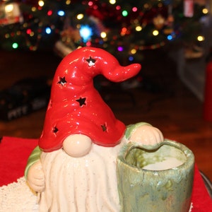 May include: A ceramic gnome figurine with a red hat and white beard. The gnome is holding a green ceramic pot with a white interior.