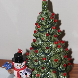 May include: A ceramic Christmas tree with red ornaments and a red star on top. Beside it is a snowman figurine wearing a black top hat and red scarf, holding a bird and birdhouse.
