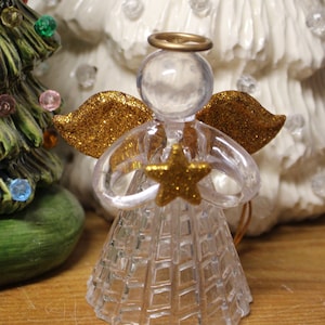 May include: A clear, faceted angel figurine with gold glitter wings and a gold star. The angel has a gold halo and is holding the star. The figurine is a Christmas decoration.