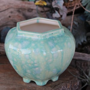 May include: A ceramic planter with a rounded body and a hexagonal opening. The planter is glazed in shades of turquoise and pale yellow, with a speckled pattern. It rests on three small feet, suitable for indoor or outdoor use.