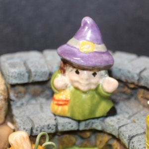 May include: A small ceramic witch figurine wearing a purple hat and green dress, holding a small orange and yellow basket. The figurine is sitting on a stone step.