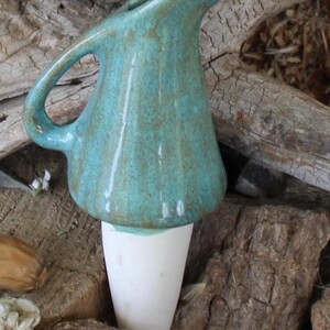 May include: A ceramic plant watering spike featuring a glossy turquoise glazed top shaped like a small pitcher with a handle and spout, attached to a tapered, unglazed white clay stake designed for insertion into soil for hydration.