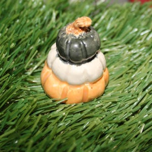 May include: A stack of three miniature ceramic pumpkins. The top pumpkin is dark green, the middle pumpkin is white, and the bottom pumpkin is orange. The pumpkins are sitting on a bed of green artificial grass.