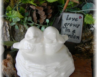 May include: White ceramic figurine of two birds perched on a nest. The nest is made of white ceramic and has a textured surface. The birds are facing each other and have their beaks open. The figurine is sitting on a wooden surface. A sign with the text "love grows in a garden" is visible in the background.
