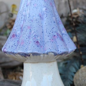 May include: A ceramic mushroom figurine with a light blue cap adorned with pink floral patterns and dark speckles. The stem is off-white with a speckled texture. The mushroom has a glossy finish and is likely a decorative item.