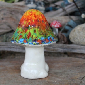 May include: A ceramic mushroom figurine with a white stem and a colorful, abstractly painted cap. The cap features a mix of red, orange, yellow, green, and blue colors.