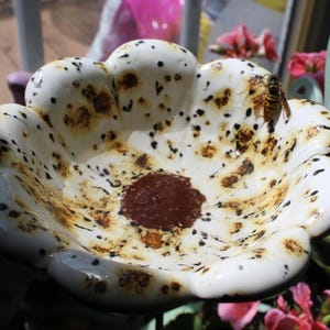 May include: A white ceramic bird bath with a flower-like shape. The bath has brown and black speckled patterns and a dark brown center. A bee rests on the edge of the bath, which is surrounded by greenery and pink flowers.