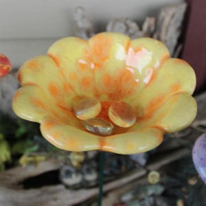 May include: A handcrafted glass flower with a yellow and orange color scheme. The flower has a petal design and contains several small, clear, and amber-colored glass stones. The flower is mounted on a green stem.