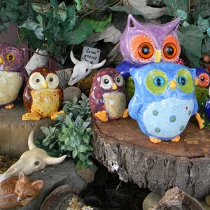 May include: A collection of colourful ceramic owl figurines, some with painted details, arranged on a wooden surface with a skull and greenery. The owls are various sizes and have different expressions. The text "love grows here" is visible on a small sign.