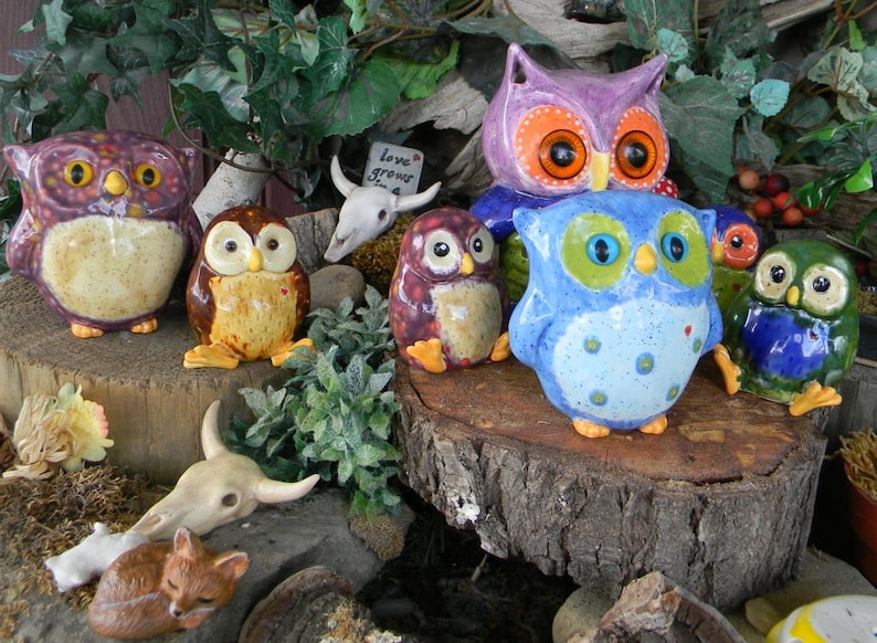 May include: A collection of colourful ceramic owl figurines, some with painted details, arranged on a wooden surface with a skull and greenery. The owls are various sizes and have different expressions. The text "love grows here" is visible on a small sign.