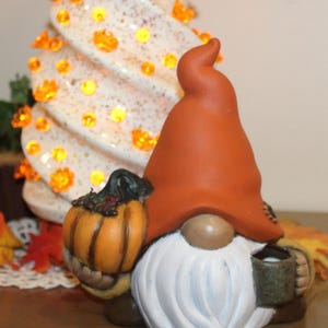 May include: A ceramic gnome figurine featuring a tall, solid orange hat, a long white beard, and a tan nose. The gnome holds a small orange pumpkin in one hand and a dark green mug in the other, with a textured surface and painted details.