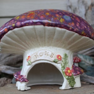 May include: A ceramic mushroom-shaped house with a glossy, mottled purple and orange cap. The cream-colored stem features a central arched opening, a banner with the embossed text HOME, and decorative sculpted green leaves and small red flowers.