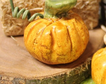May include: A small ceramic pumpkin with a green stem and a brown, wooden base. The pumpkin is painted in a light orange color and has a textured surface.