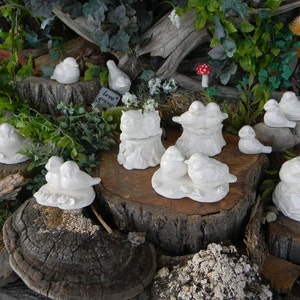 May include: A collection of white ceramic bird figurines, some in pairs, perched on tree stumps and other natural bases. The birds are arranged in various poses, some with their wings spread, others with their beaks open. The figurines are all white and have a smooth, glossy finish. The background is a natural setting with green plants and brown wood.