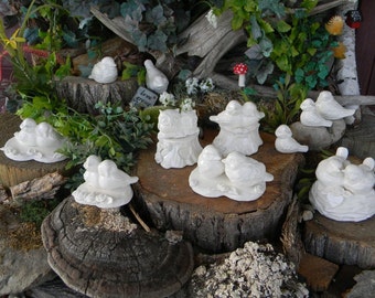 May include: A collection of white ceramic bird figurines, some in pairs, perched on tree stumps and other natural bases. The birds are arranged in various poses, some with their wings spread, others with their beaks open. The figurines are all white and have a smooth, glossy finish. The background is a natural setting with green plants and brown wood.