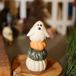 May include: A white ghost figurine with a black face sits atop a stack of three miniature pumpkins. The pumpkins are orange, green, and white. The figurine is sitting on a wooden slice.