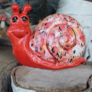 May include: A whimsical, red ceramic snail figurine with a smiling face and expressive eyes. The snail's shell features a colorful, speckled pattern in shades of red, orange, and black. The figurine is a decorative item, likely for a garden or home decor.
